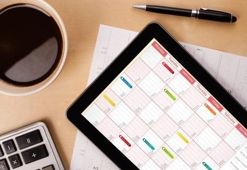 image portraying how to create a schedule on a tablet with online monthly calendar view pls a calculator and black coffee and a black pen on a desk.