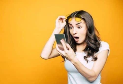 Woman wearing a white tshirt, yellow sunglasses held above her eyebrows and a silver smartphone with a surprised look on her face at how much she can earn from pictures of her feet on a bright yellow background.