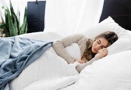 Woman laying down in bed covered by bed sheets doing get paid to sleep side job